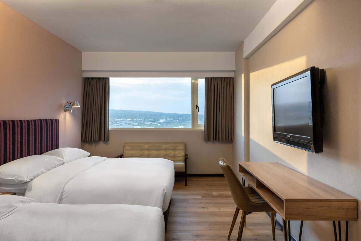 A hotel room with two beds, a desk, a chair, a TV, and a window with a view of the sea.