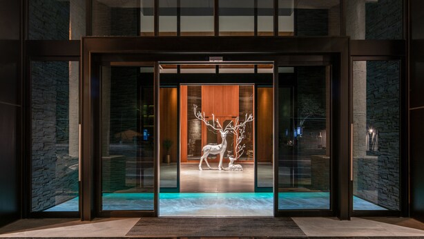 Una entrada de edificio moderna con puertas de vidrio, interior de madera y una escultura de ciervo.