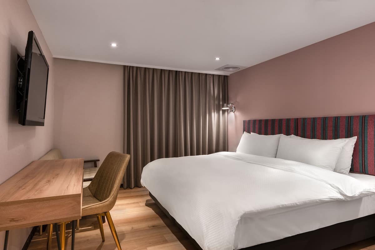 A hotel room with a wooden desk, a bed with white linens, a television mounted on the wall, and a striped headboard.