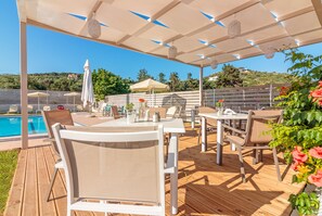 Un patio al aire libre con mesas y sillas, piscina y una pérgola con luces colgantes.