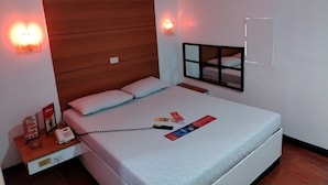 Une chambre d’hôtel avec une tête de lit en bois, un lit avec un matelas blanc, une table de chevet avec un téléphone et un menu, et un appareil de climatisation fixé au mur.