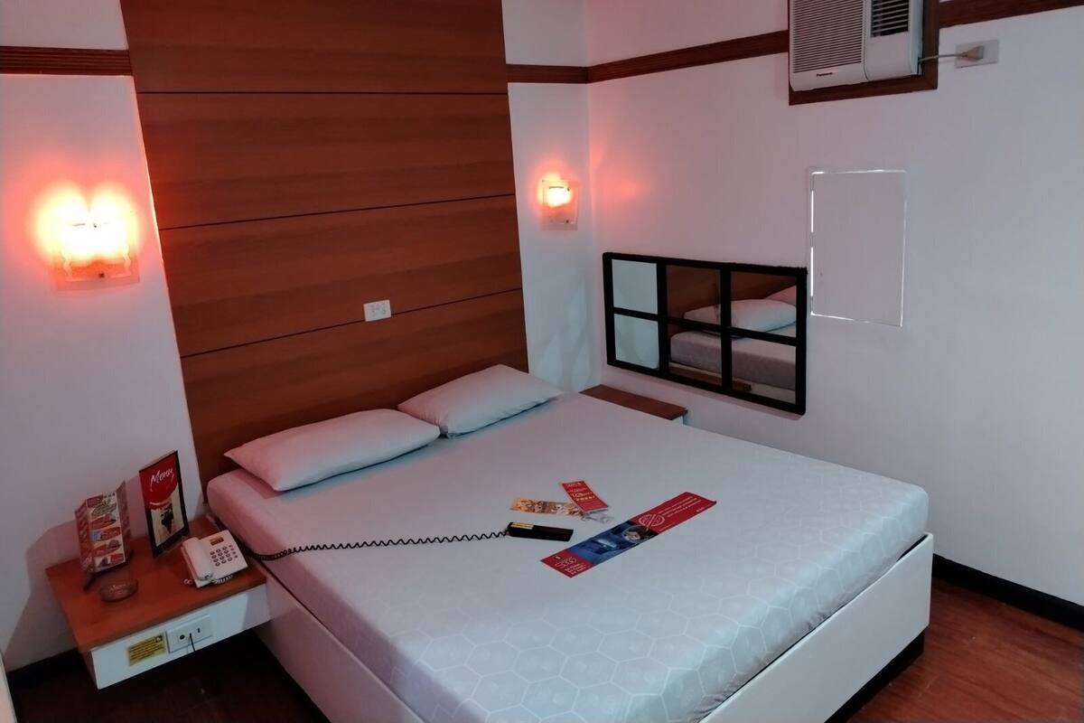 A hotel room with a wooden headboard, a bed with a white mattress, a bedside table with a phone and a menu, and an air conditioning unit on the wall.
