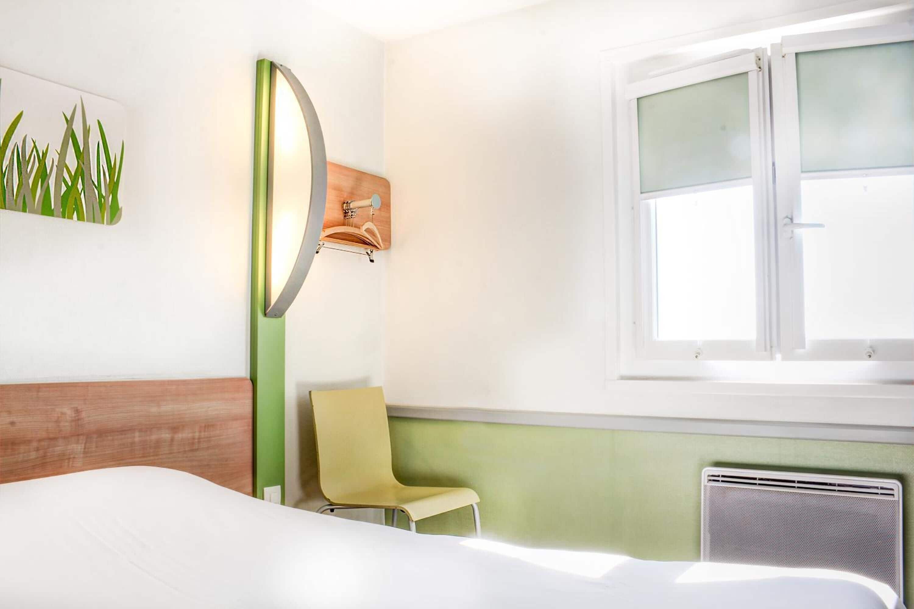 A hotel room with a bed, a chair, a wall-mounted mirror, and a window with blinds.
