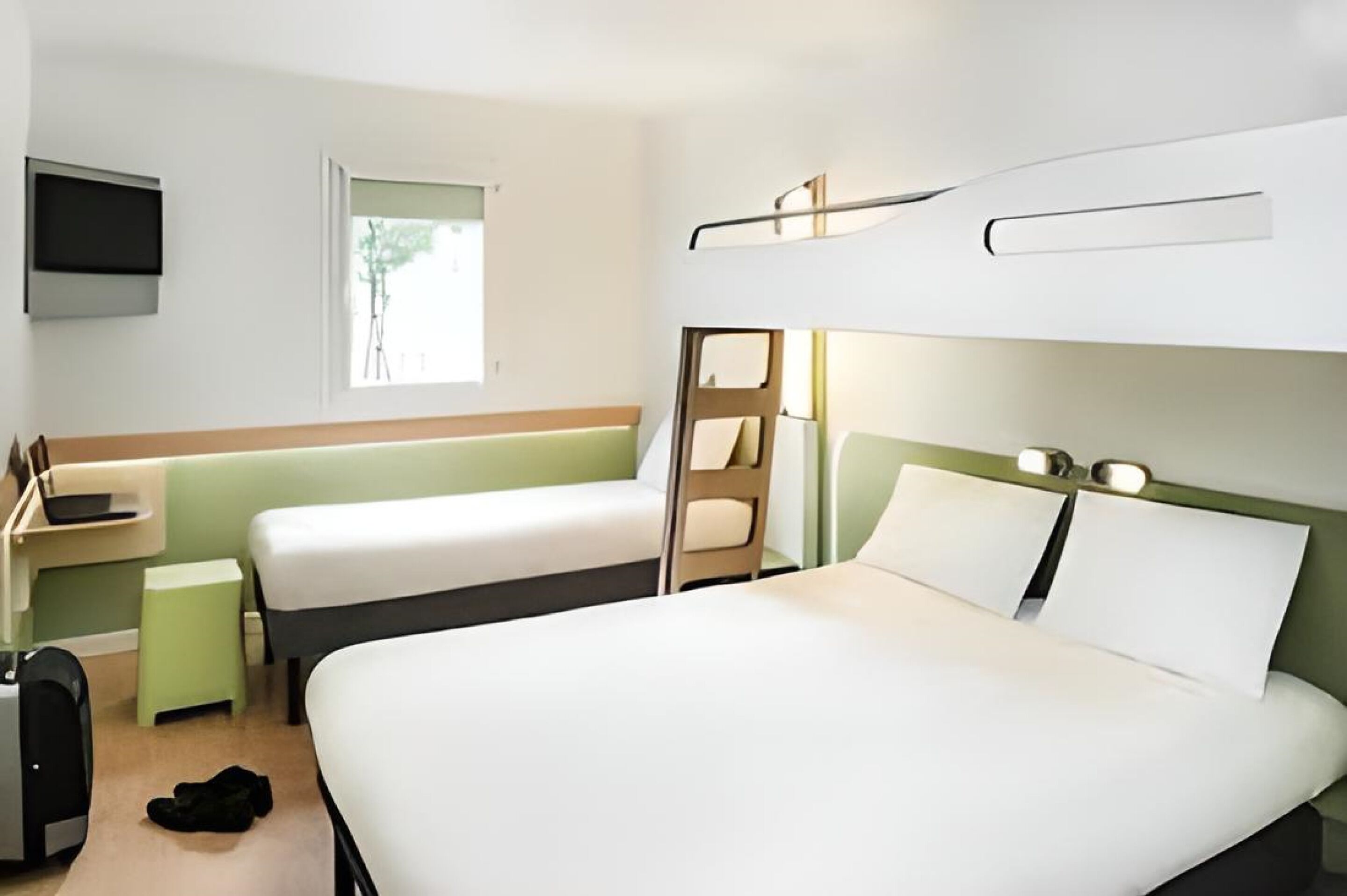 A hotel room with a large bed, two single beds, a TV mounted on the wall, and a window providing natural light.