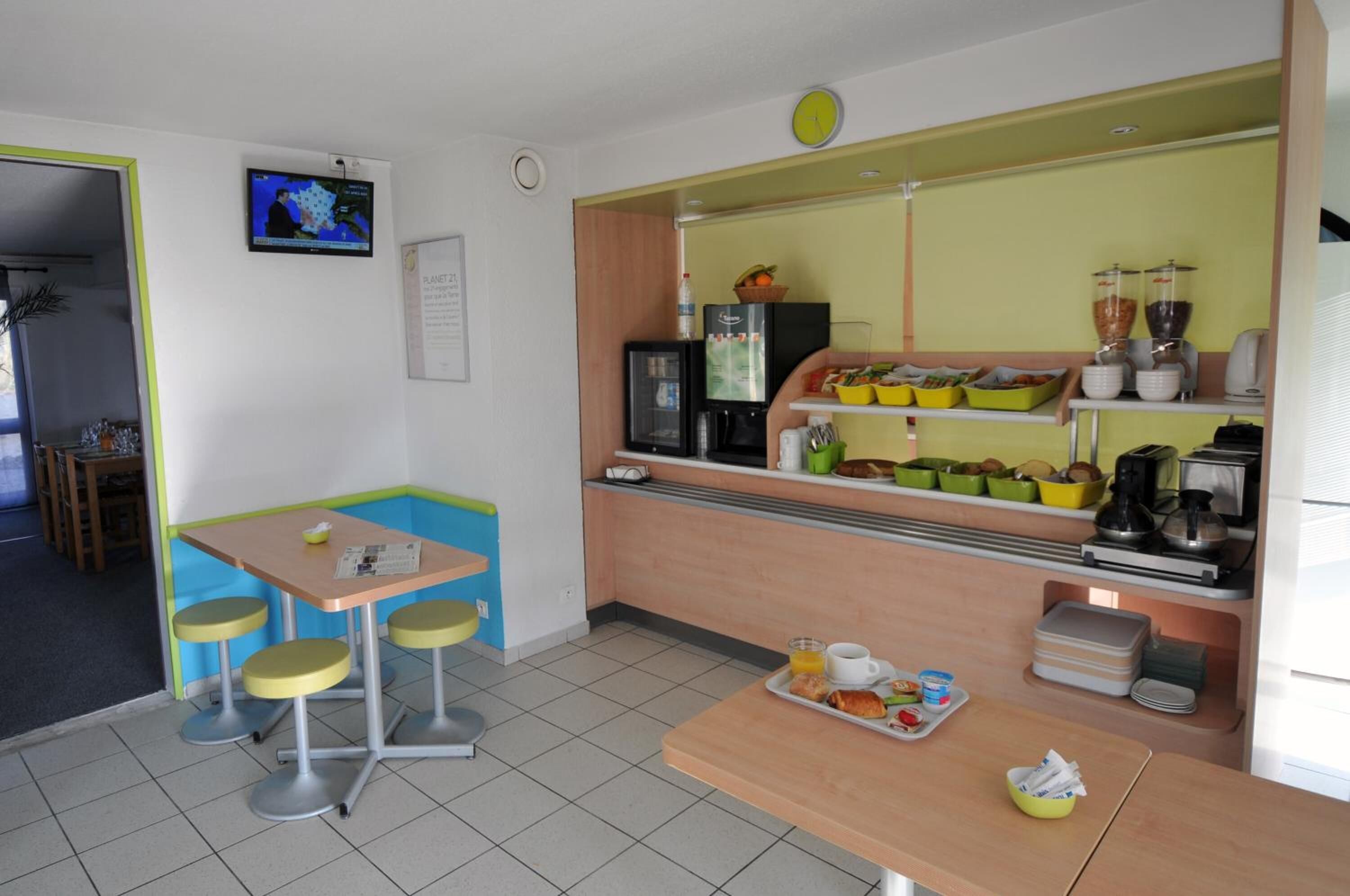 A small, brightly lit room with a kitchenette, a table with chairs, and a television on the wall.