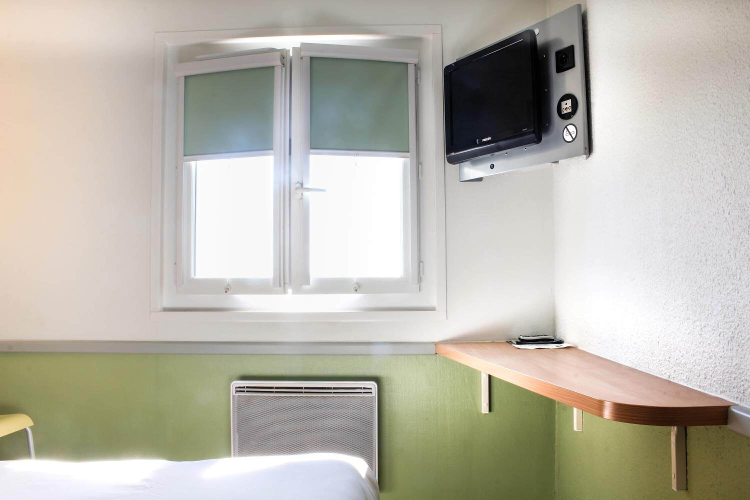 Une chambre d’hôtel avec un lit, une fenêtre avec des stores, un téléviseur fixé au mur et une petite étagère en bois.