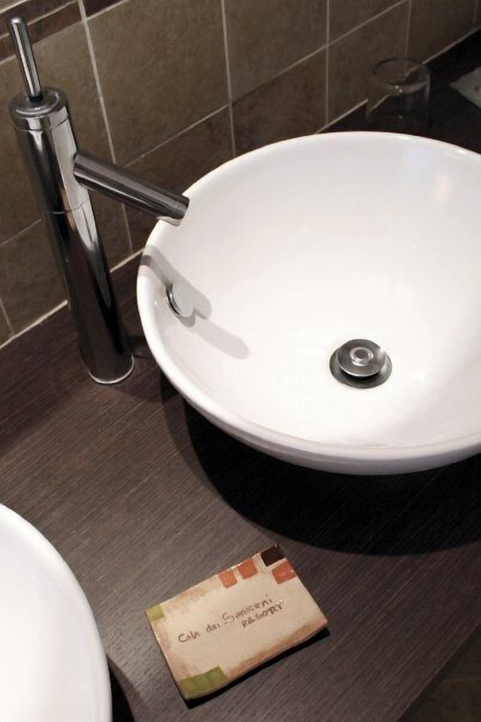 Un bagno con un lavandino bianco, un rubinetto cromato e un piccolo biglietto sul ripiano.