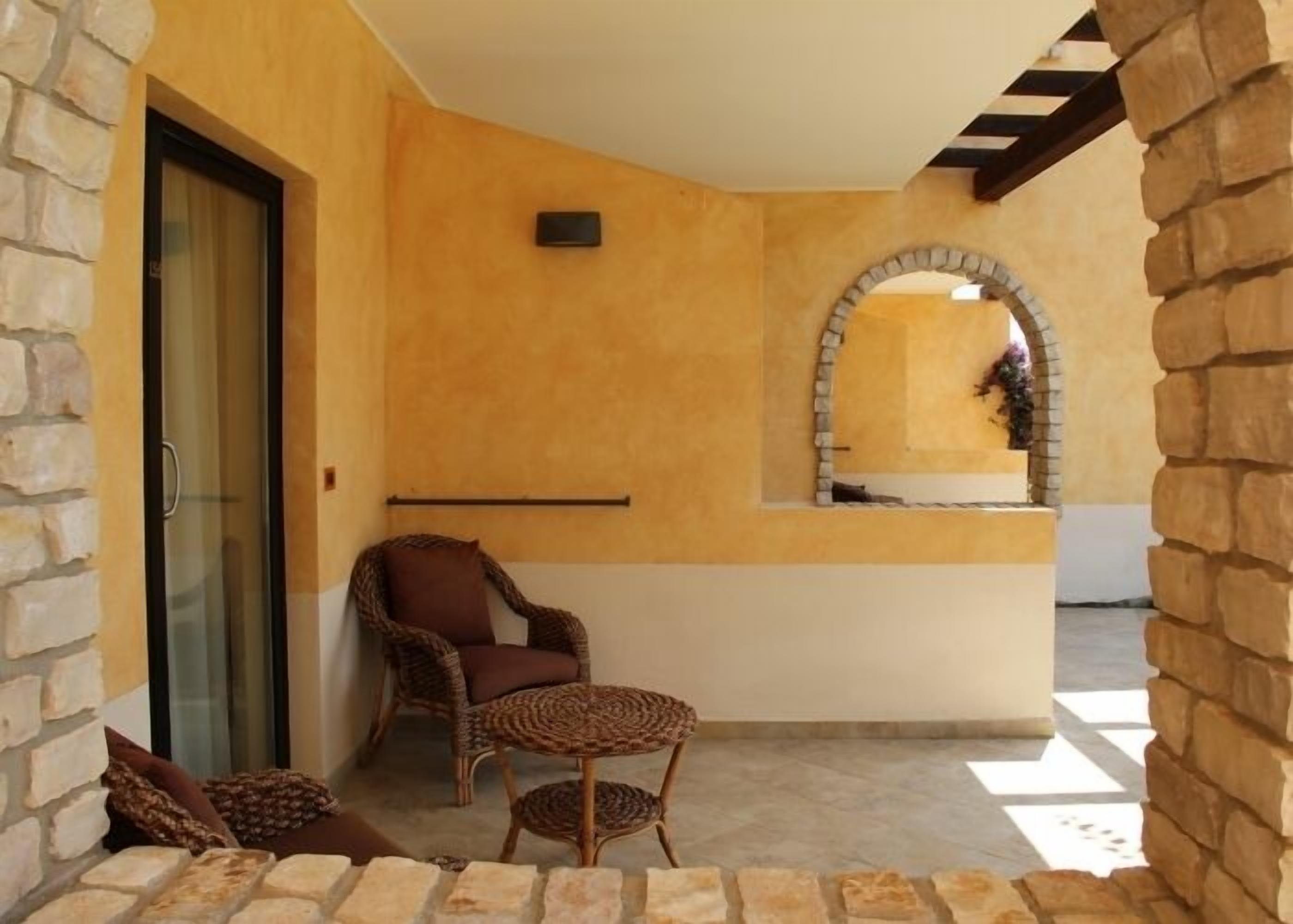 A cozy outdoor seating area with a wicker chair and a small round table, a glass door, and a stone wall.