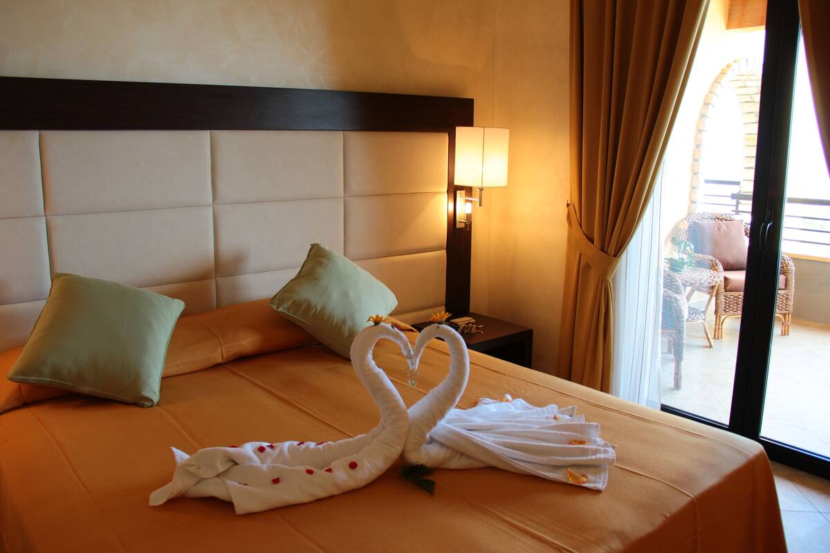 A hotel room with a bed, pillows, a nightstand with a lamp, and a view of the outside.