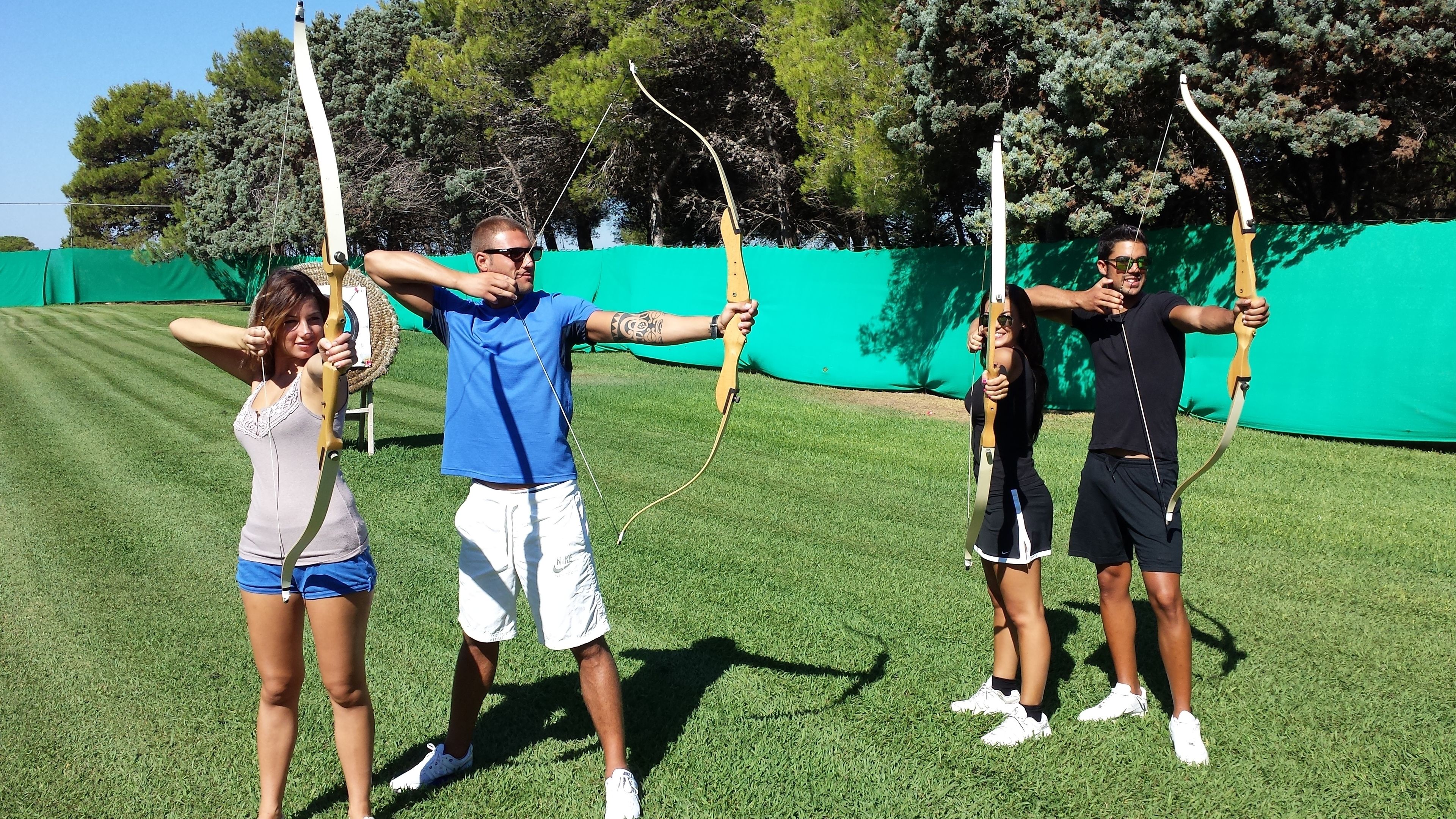 Four people holding bows on a grassy field with a green fence in the background.