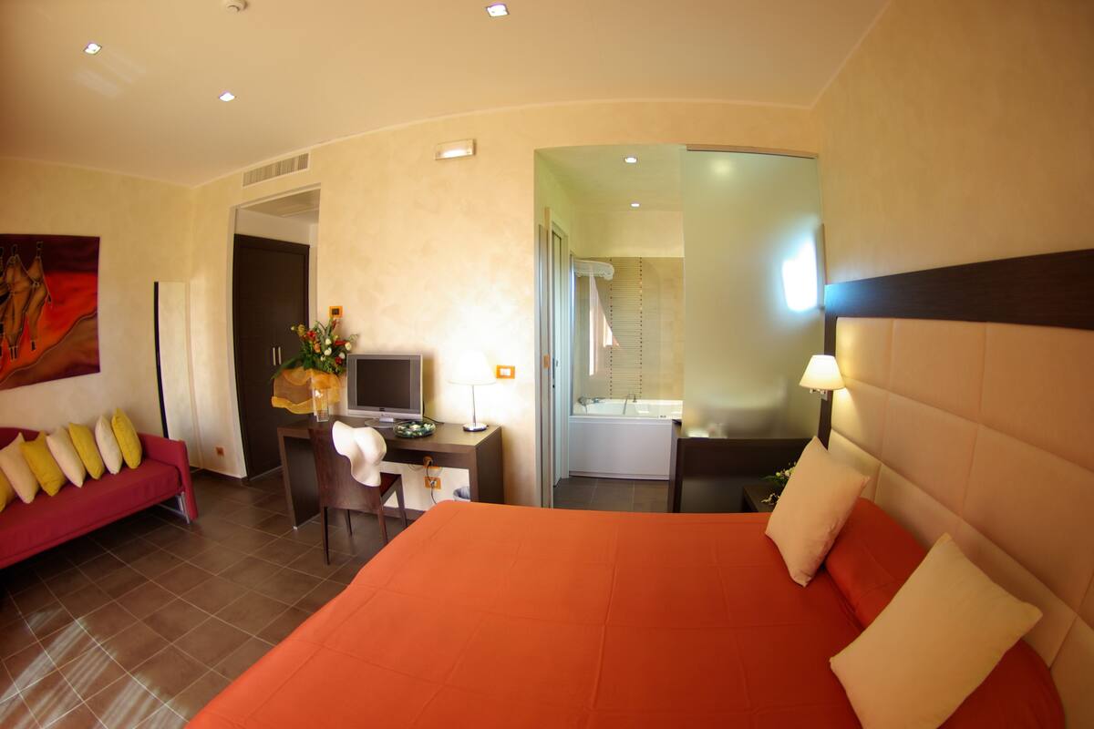 A hotel room with a large bed, a desk with a computer, a TV, and a bathroom visible through an open door.