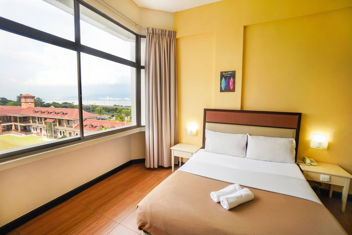 A hotel room with a large window, a bed with a brown bedspread, two bedside tables with lamps, and a view of a building with a clock tower.