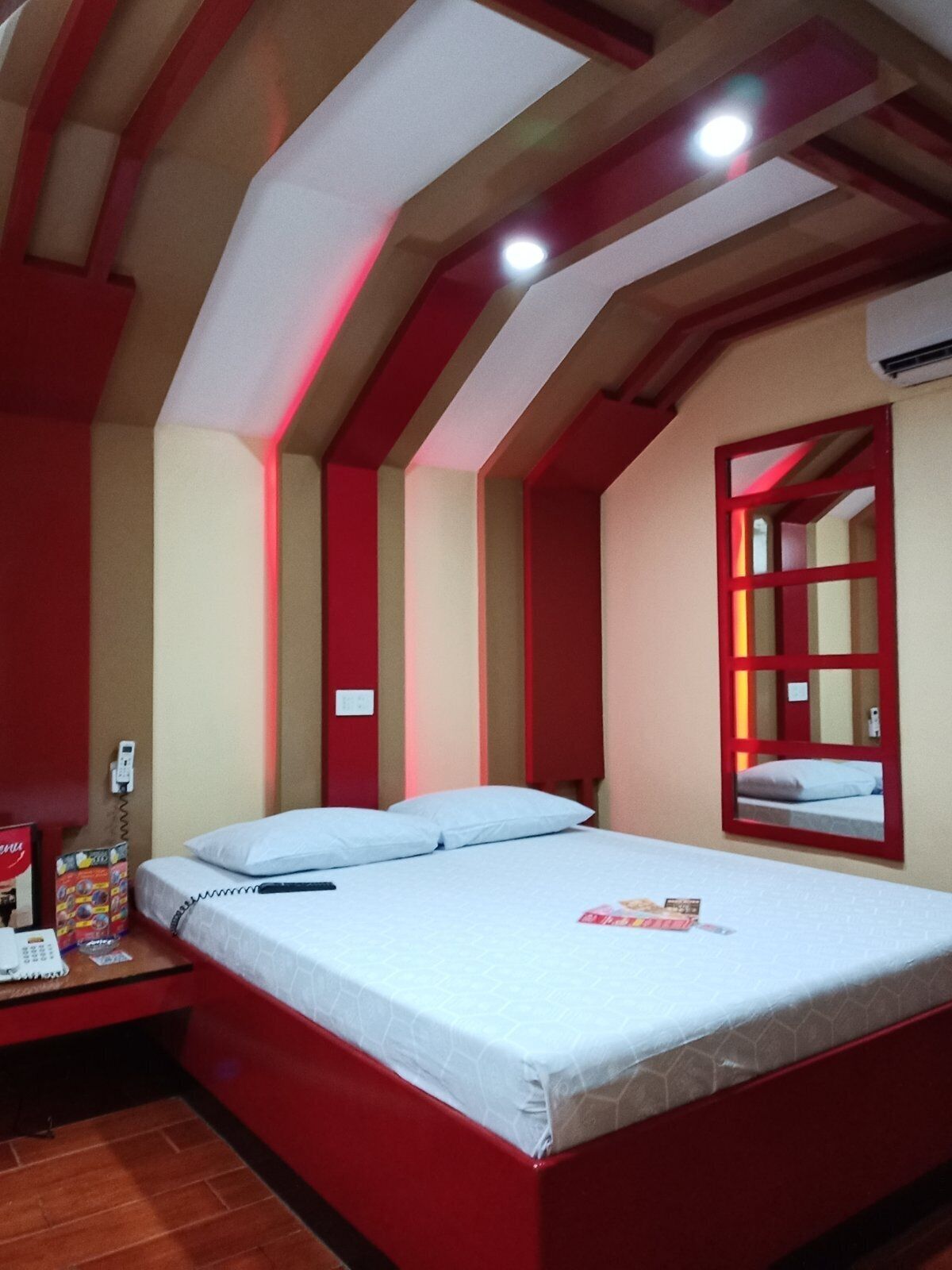 Quarto de hotel com cama, mesa de cabeceira, espelho e uma decoração em tons de vermelho e bege.