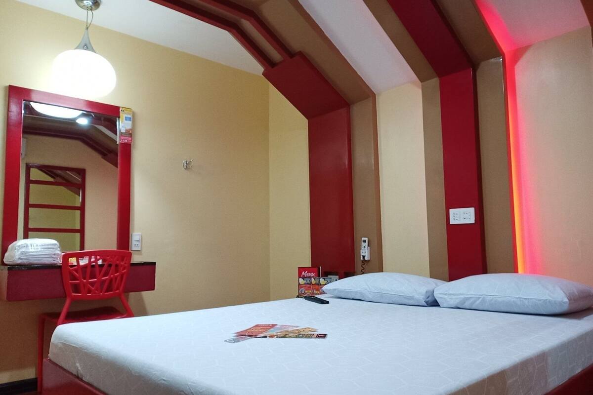 Ein Einzelbett mit weißer Matratze und Kissen, ein roter Sessel, ein kleiner Schreibtisch mit Spiegel und eine Wandleuchte.