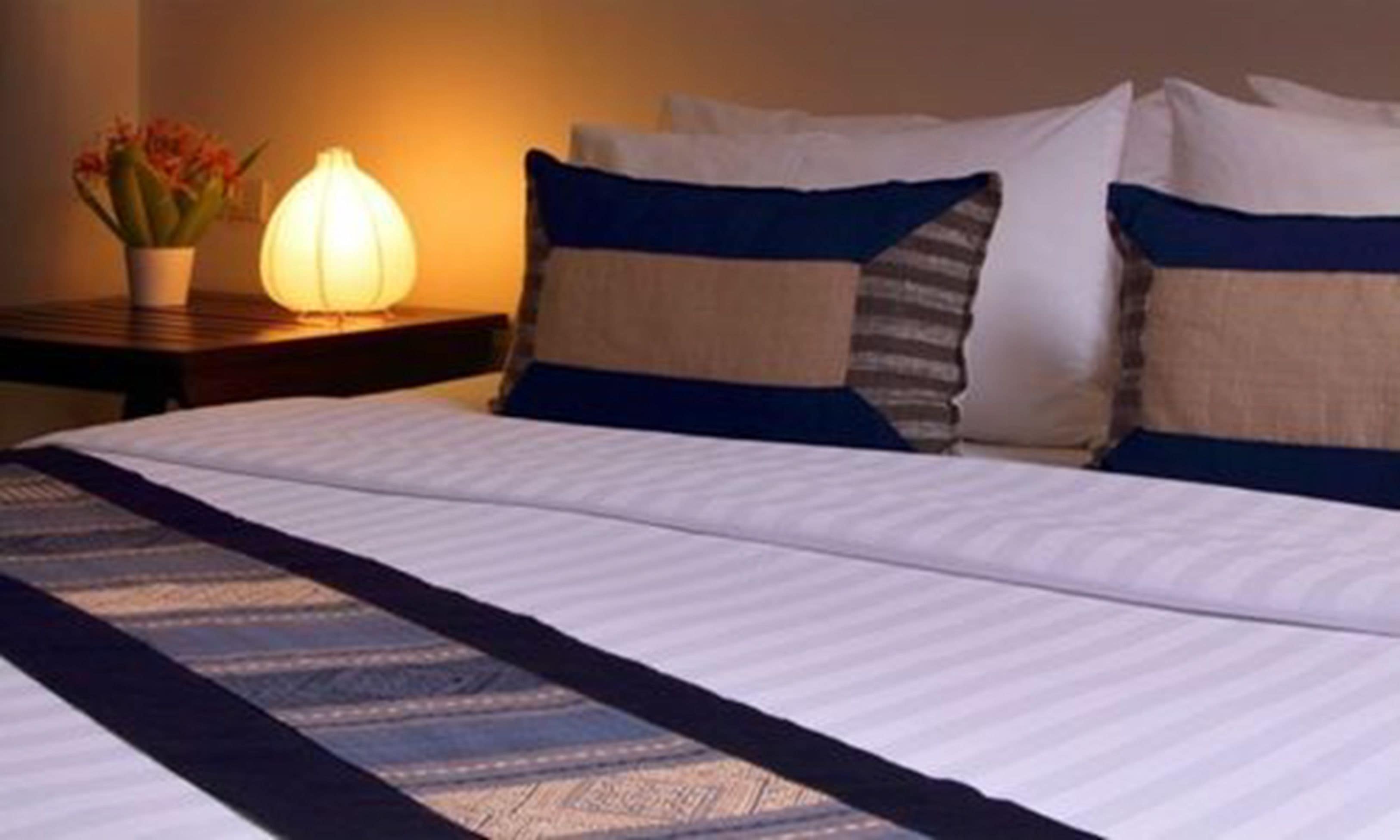 A neatly made bed with a striped bedspread and pillows, a bedside lamp, and a vase with flowers on a nightstand.