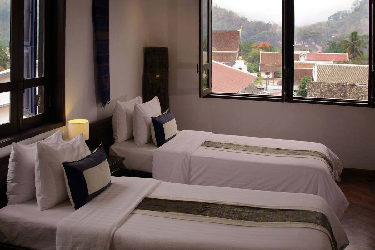 Habitación de hotel con dos camas, una mesita de noche, una lámpara y un ventanal con vistas a edificios y colinas.