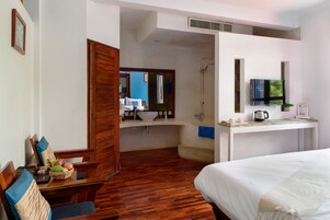 A hotel room with a wooden floor, a bed, a desk with a television, a shower, and a dining area visible through an open door.