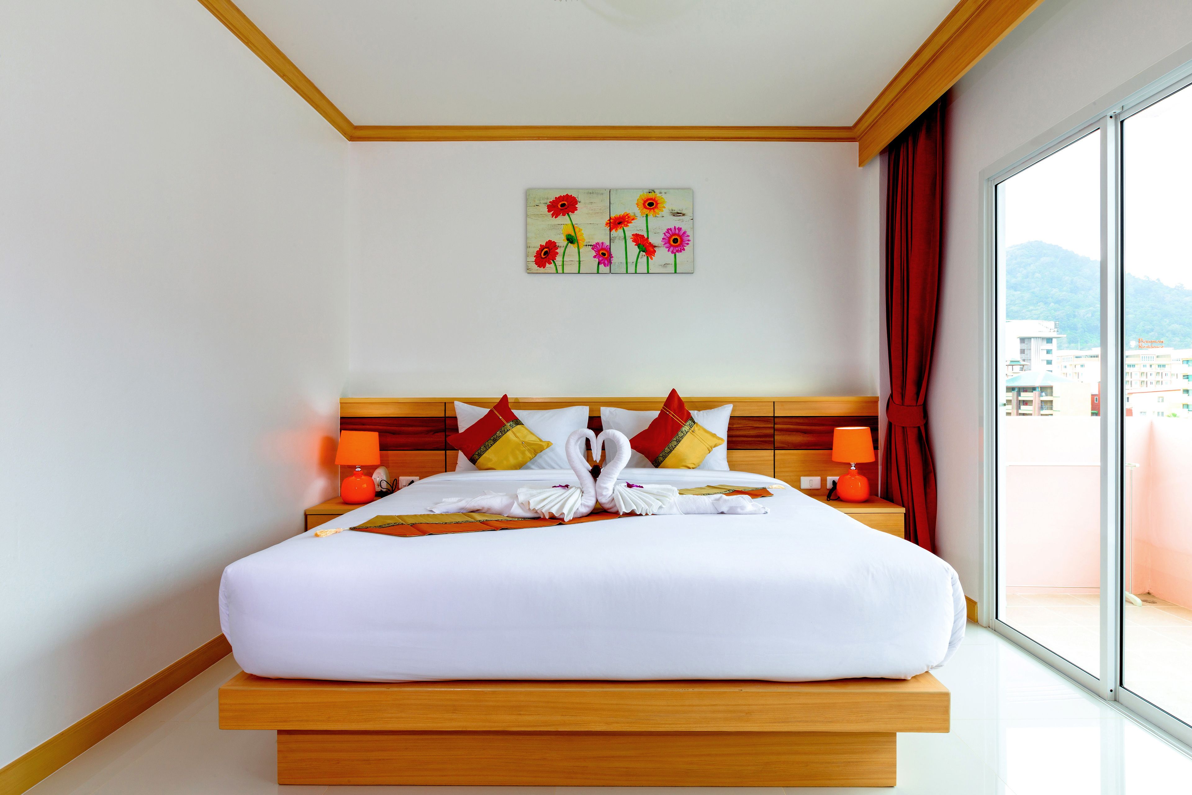 A hotel room with a wooden bed, white bedding, a painting of flowers on the wall, and a view of buildings through the window.