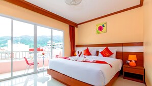 Habitación de hotel con una cama grande, una mesita de noche con lámpara, vistas de la ciudad a través de amplios ventanales y un balcón con una silla roja.