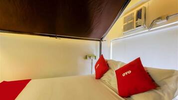 A single bed with red pillows and a white mattress, a wall-mounted air vent, and a partial view of a window.