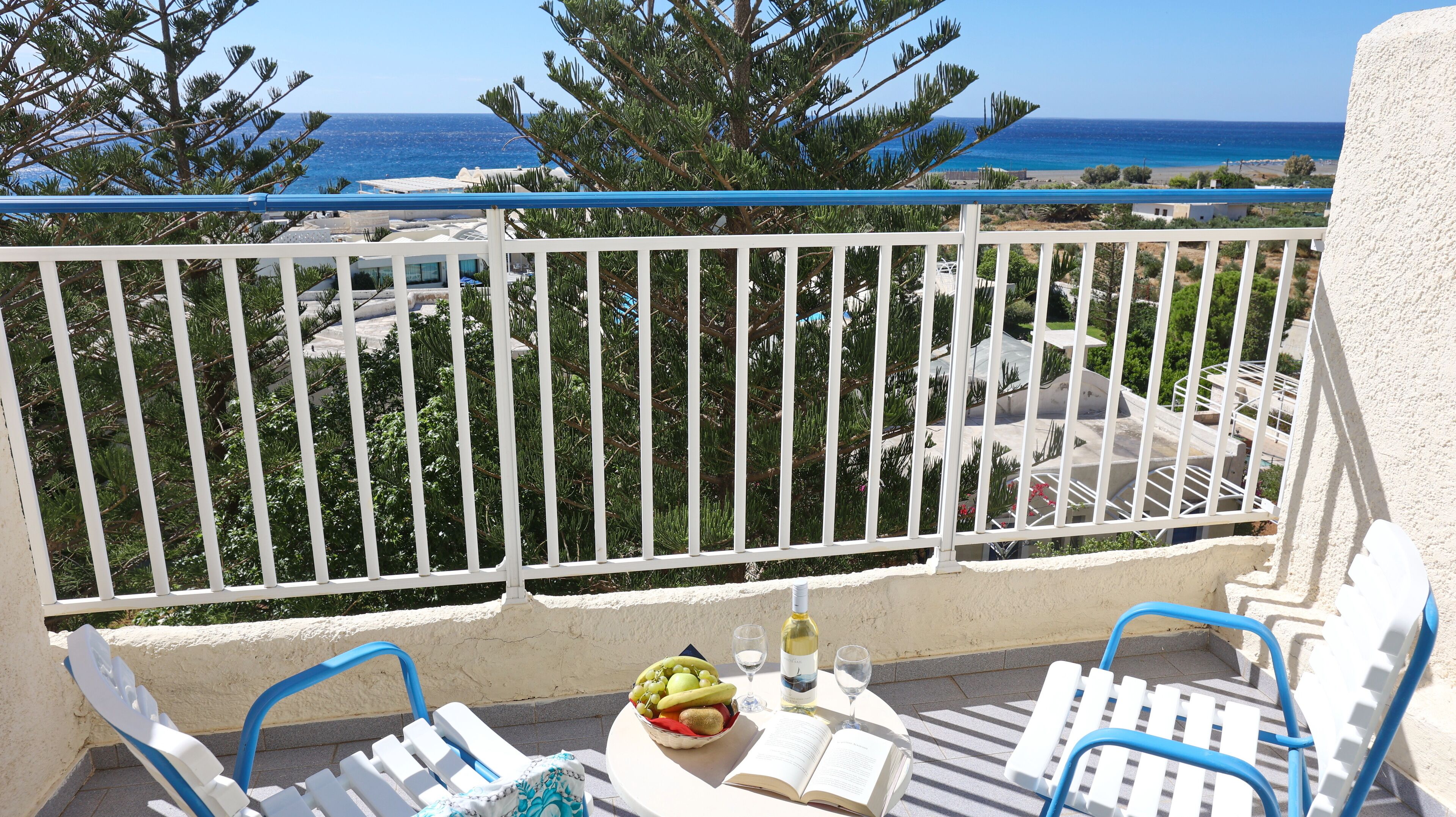 Un balcone con vista sul mare, ringhiere bianche e un tavolo con un libro, bicchieri e una ciotola di frutta.