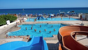 A pool area with multiple swimming pools, a water slide, and beach umbrellas. - Mare Blue Hotel (Ierapetra)