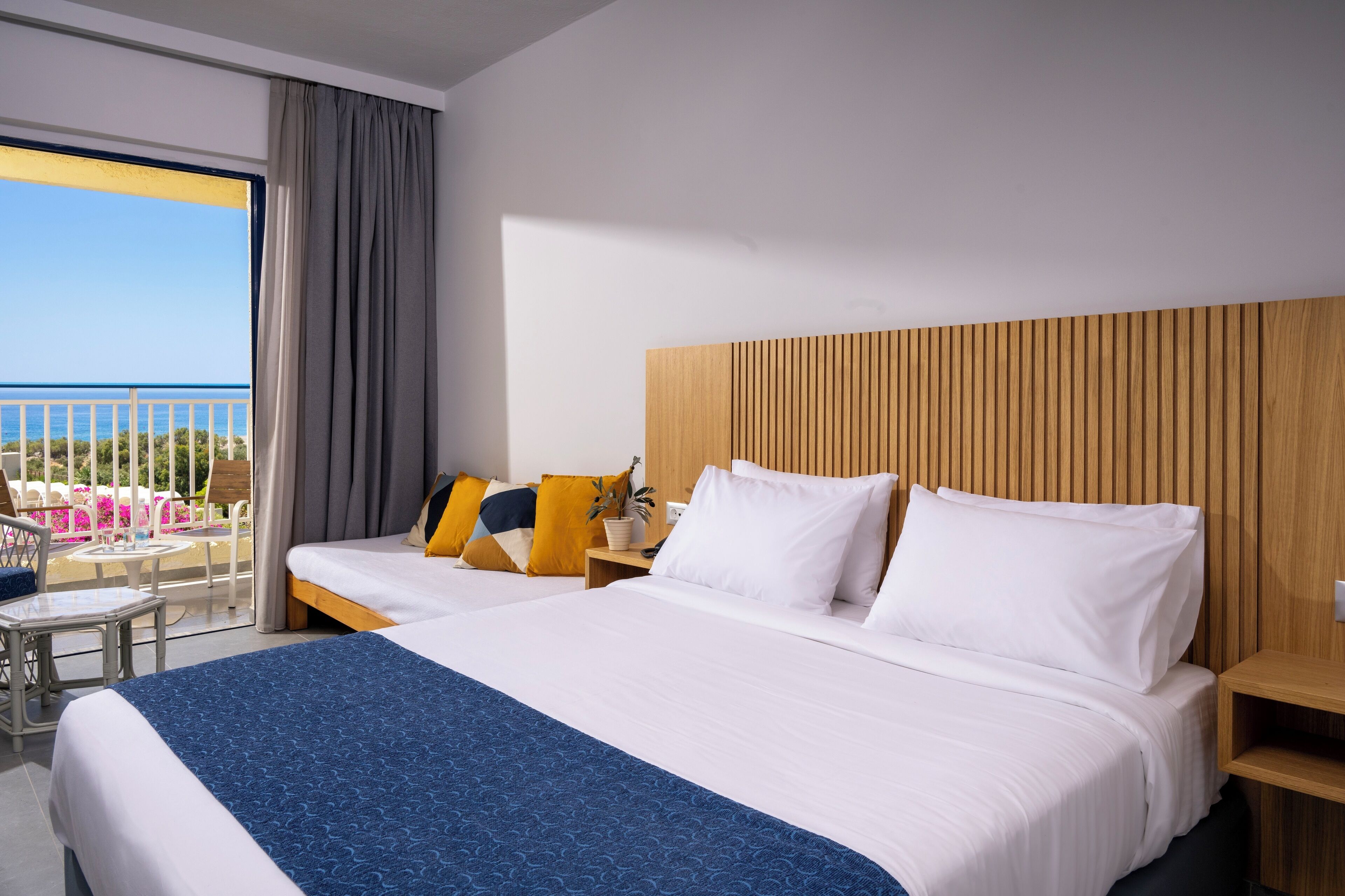 A hotel room with a large bed, two smaller beds, a balcony with a view of the sea, and a wooden headboard.