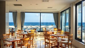 A restaurant with tables and chairs, large windows, and a view of the sea. - Mare Blue Hotel (Ierapetra)