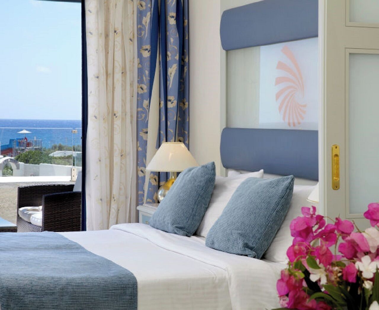 A hotel room with a bed, a nightstand, a lamp, and a view of the sea.
