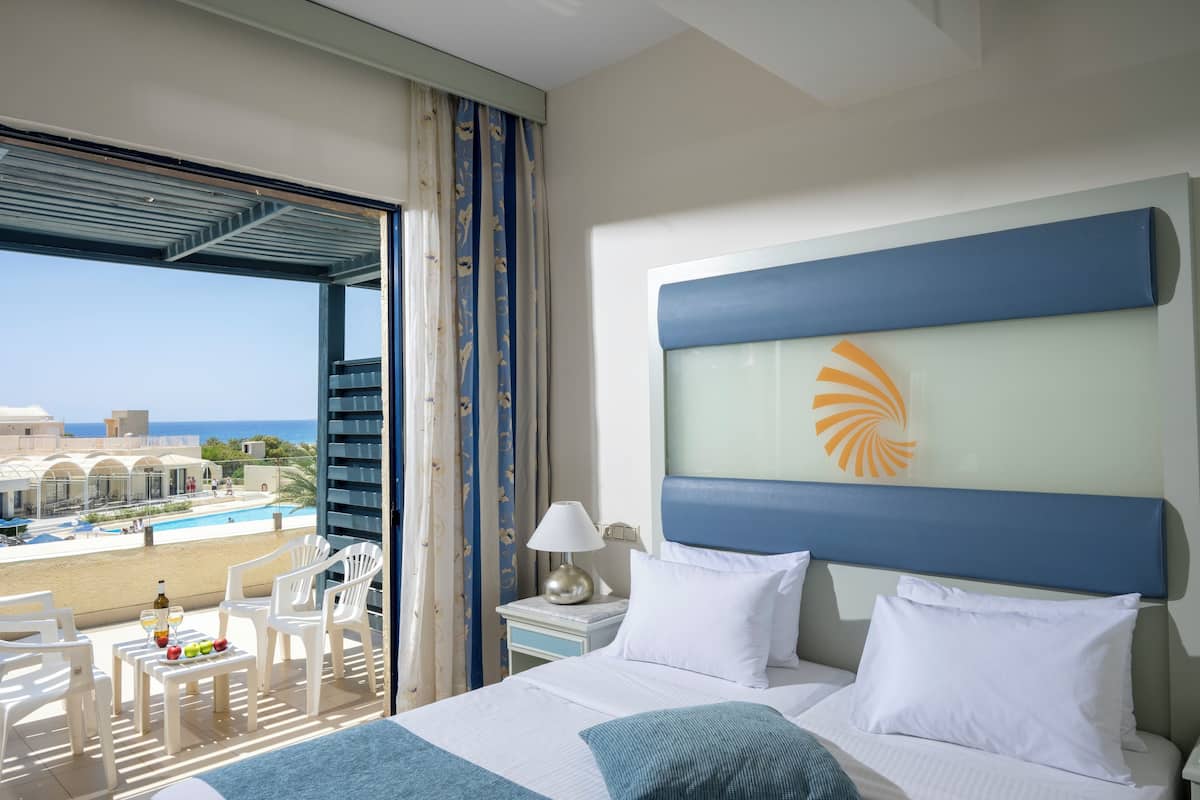 A hotel room with a bed, a nightstand, a lamp, and a view of a pool and beach.
