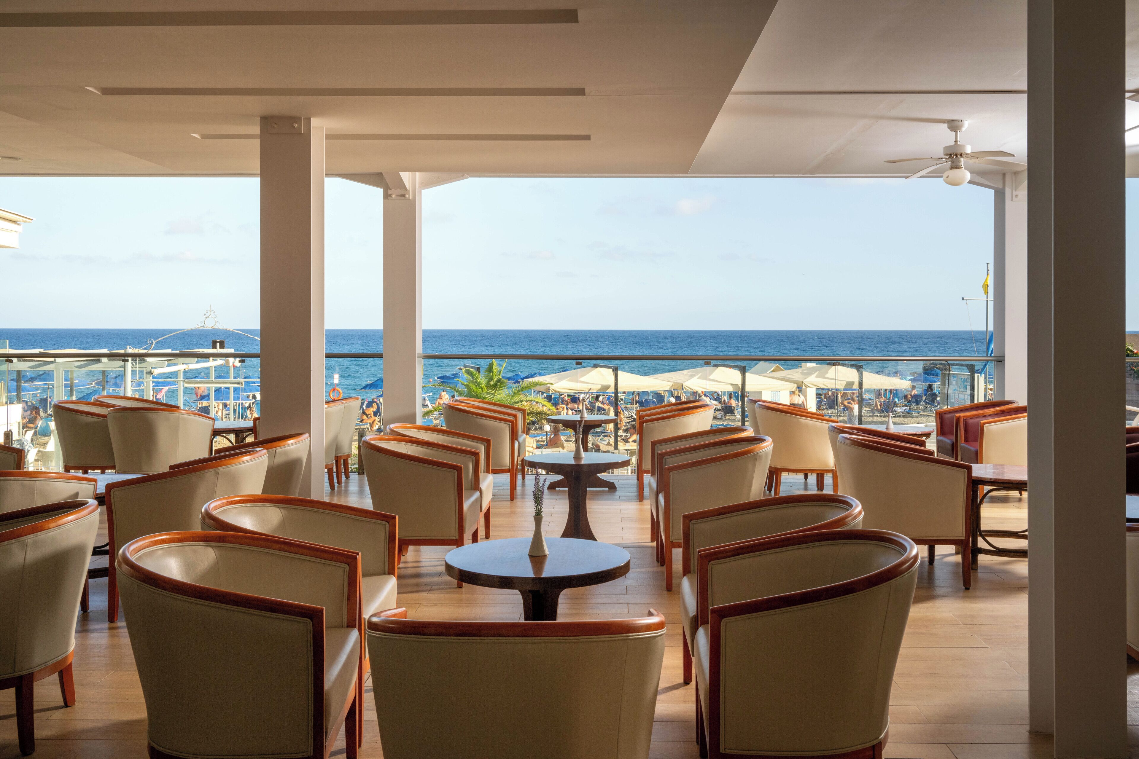 Un espace extérieur ensoleillé, aménagé avec des tables et des chaises, offrant une vue sur une plage et l’océan.