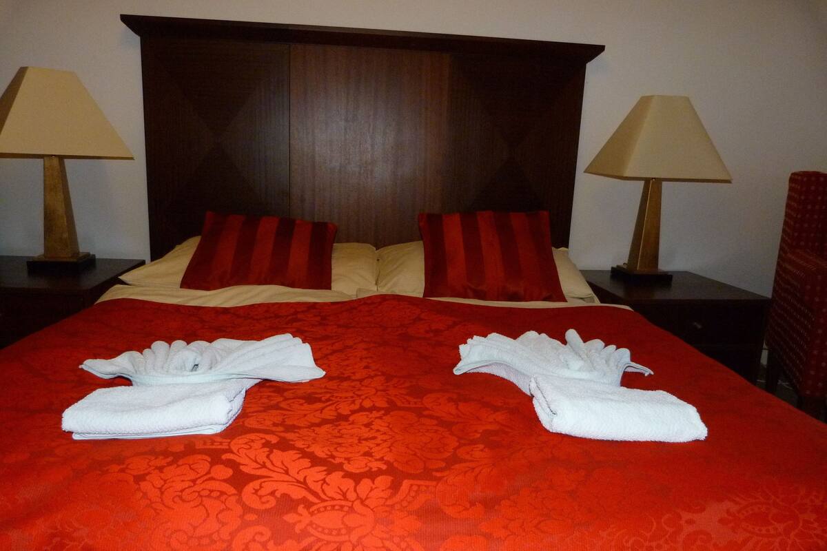 A hotel room with a large bed, two bedside lamps, and a red and beige bedspread.