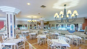 Un comedor amplio con mesas y sillas blancas, una lámpara de araña en el centro y una barra de buffet al fondo.