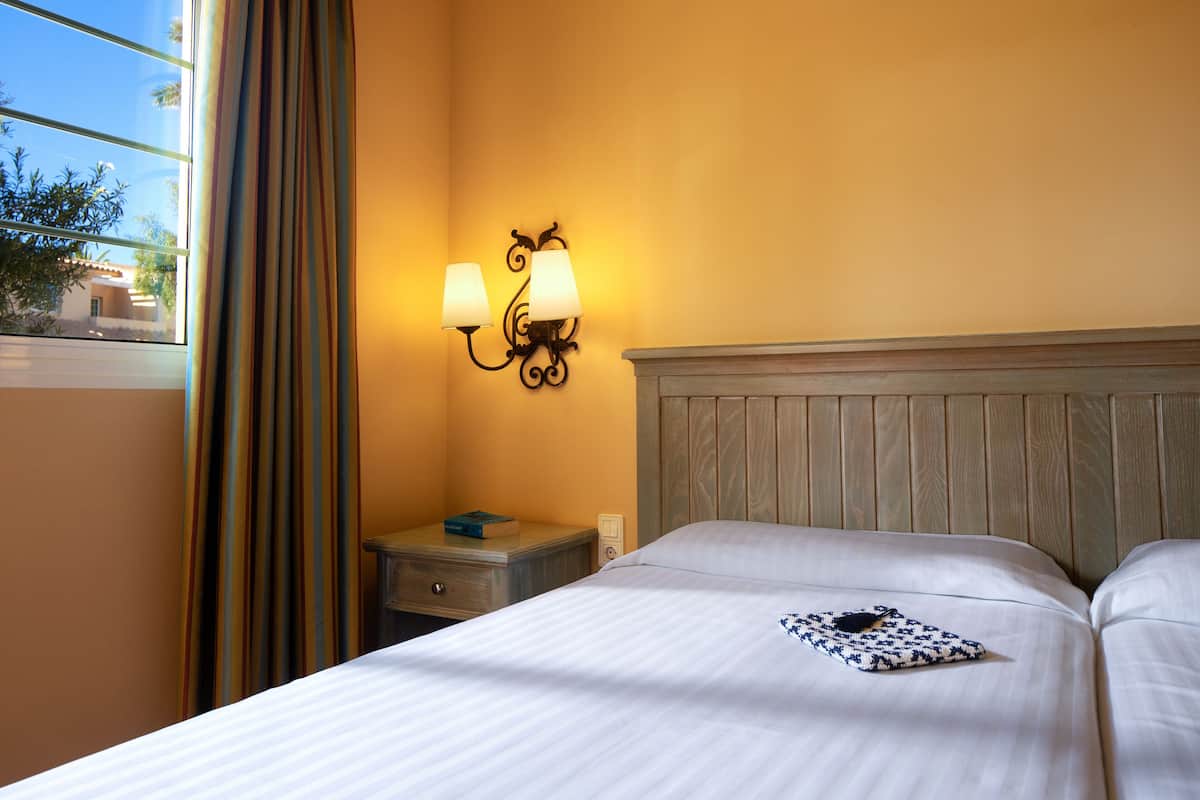 Una habitación de hotel con cama, mesita de noche, lámpara de pared y ventana con cortinas.