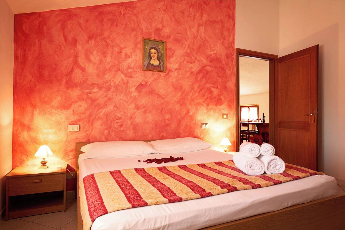 A hotel room with a large bed, a nightstand with a lamp, a wooden door, and a framed picture on the wall.