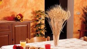 Een eettafel met een wit tafelkleed, een vaas met gedroogde planten, een dienblad met croissants en een glas rode sappen, en een glas sinaasappelsap.