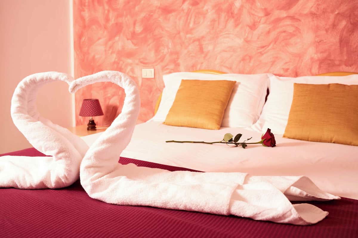 A hotel room with a bed, a heart-shaped towel arrangement, a rose, and a lamp.
