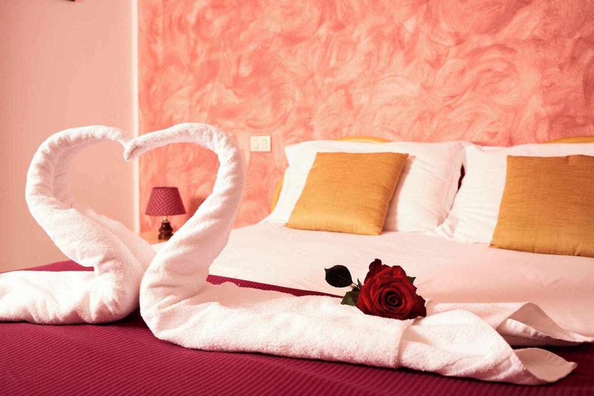 A bed with white and yellow pillows, a red rose, and two towels arranged in a heart shape.