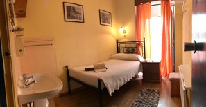 A small, simple bedroom with a single bed, a bedside table, a nightstand, a lamp, a window with curtains, and a bathroom with a sink and toilet. - Pensão Residencial Flor dos Cavaleiros (Lisbon)