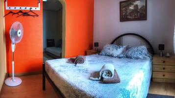 Une chambre à coucher avec un lit, une table de chevet, une commode, un ventilateur et un tableau accroché au mur.