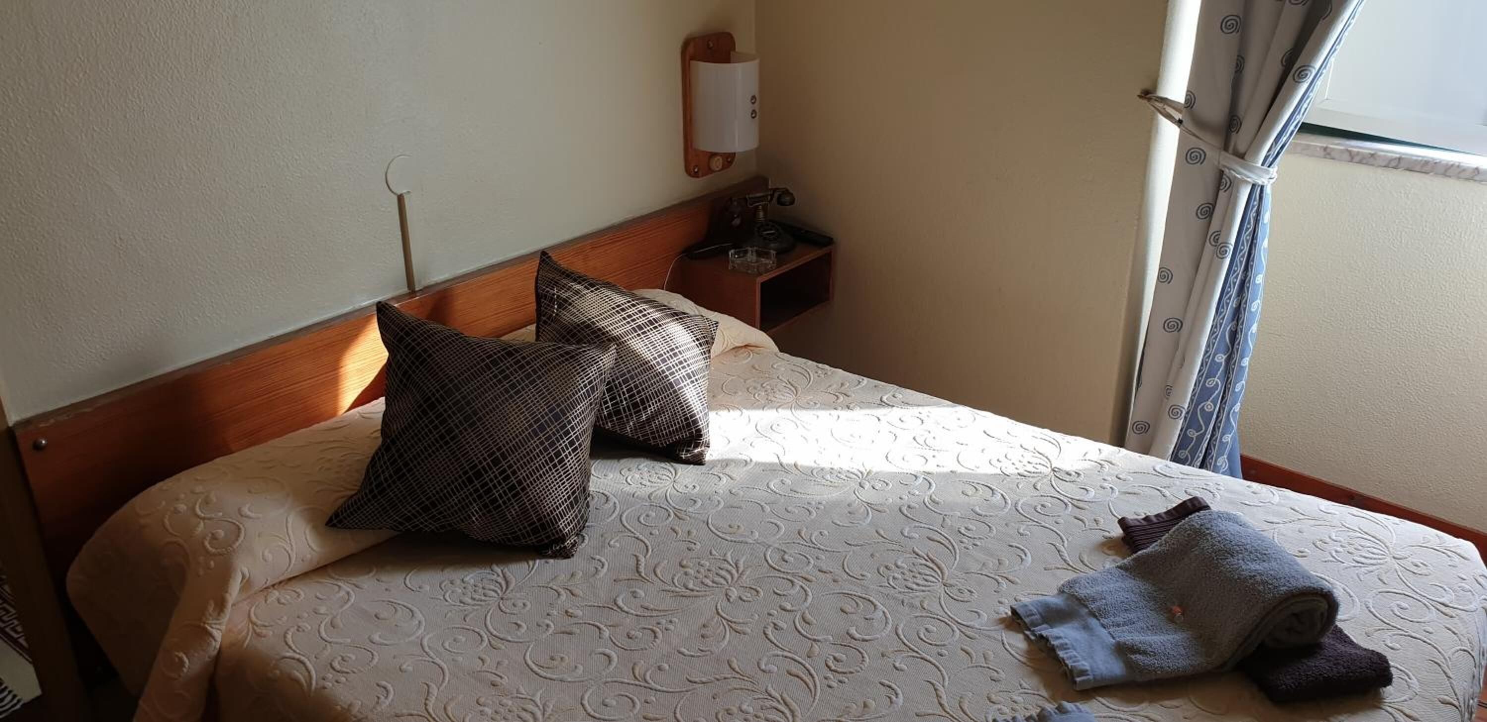 Una cama bien tendida con ropa de cama estampada, dos almohadas, una mesita de noche con lámpara y una ventana con cortinas.