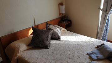 Una cama bien tendida con ropa de cama estampada, dos almohadas, una mesita de noche con lámpara y una ventana con cortinas.