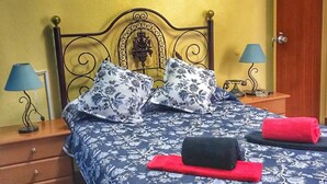 Un lit recouvert d’une couvre-lit à motifs floraux, deux oreillers assortis, une serviette rouge et une serviette noire. Une table de chevet en bois, sur laquelle se trouve une lampe, est placée de chaque côté du lit. Un tableau est accroché au mur, au-dessus du lit.