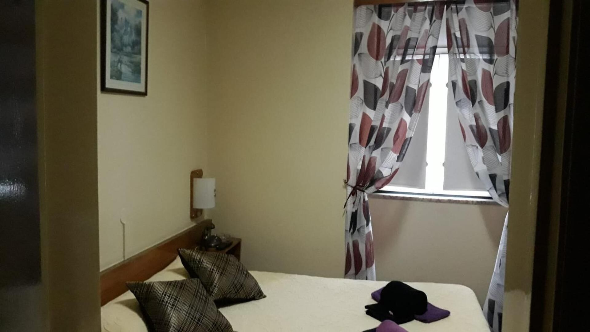 A hotel room with a bed, two pillows, a nightstand with a lamp, and a window with patterned curtains.