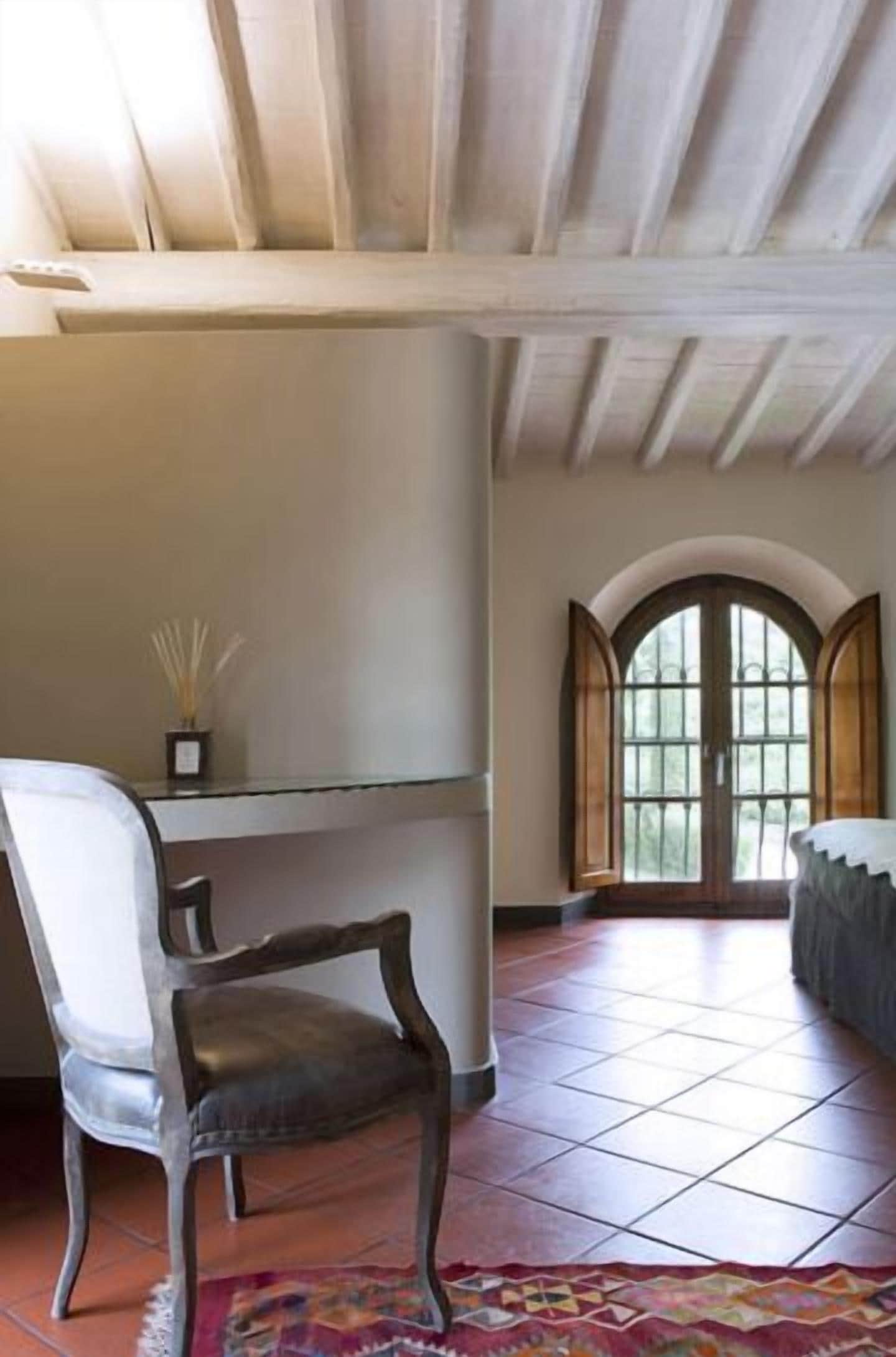 A room with a wooden ceiling, a chair, a table, and a window with arched top.