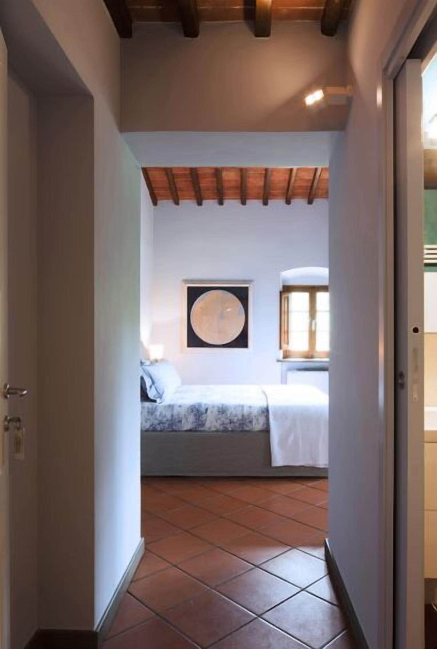 Ein schmaler Flur führt zu einem Schlafzimmer mit einem Bett, einem großen, runden Kunstwerk an der Wand und einem Fenster mit Jalousien.