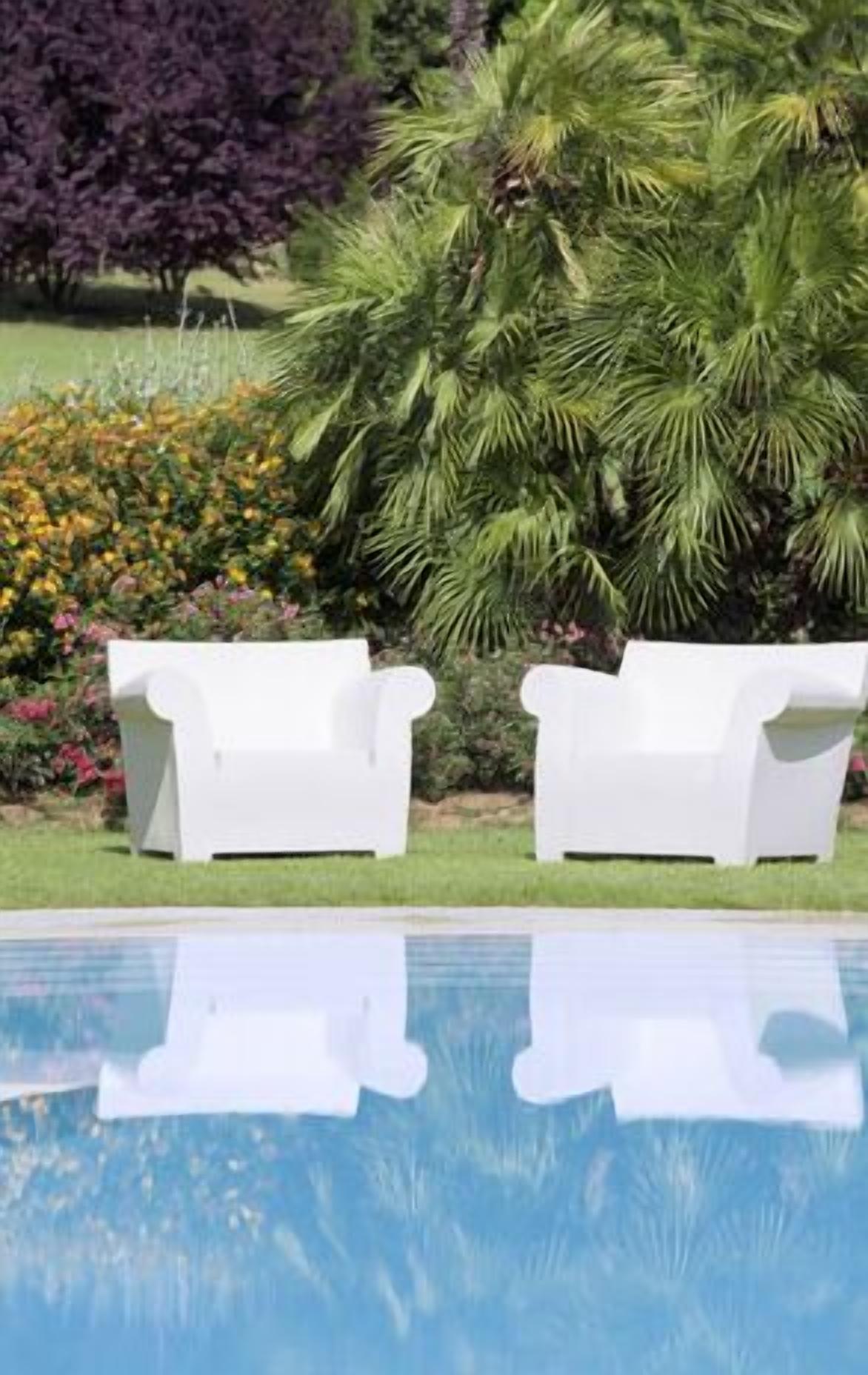 A pool with two white lounge chairs facing it, surrounded by lush greenery and flowers.