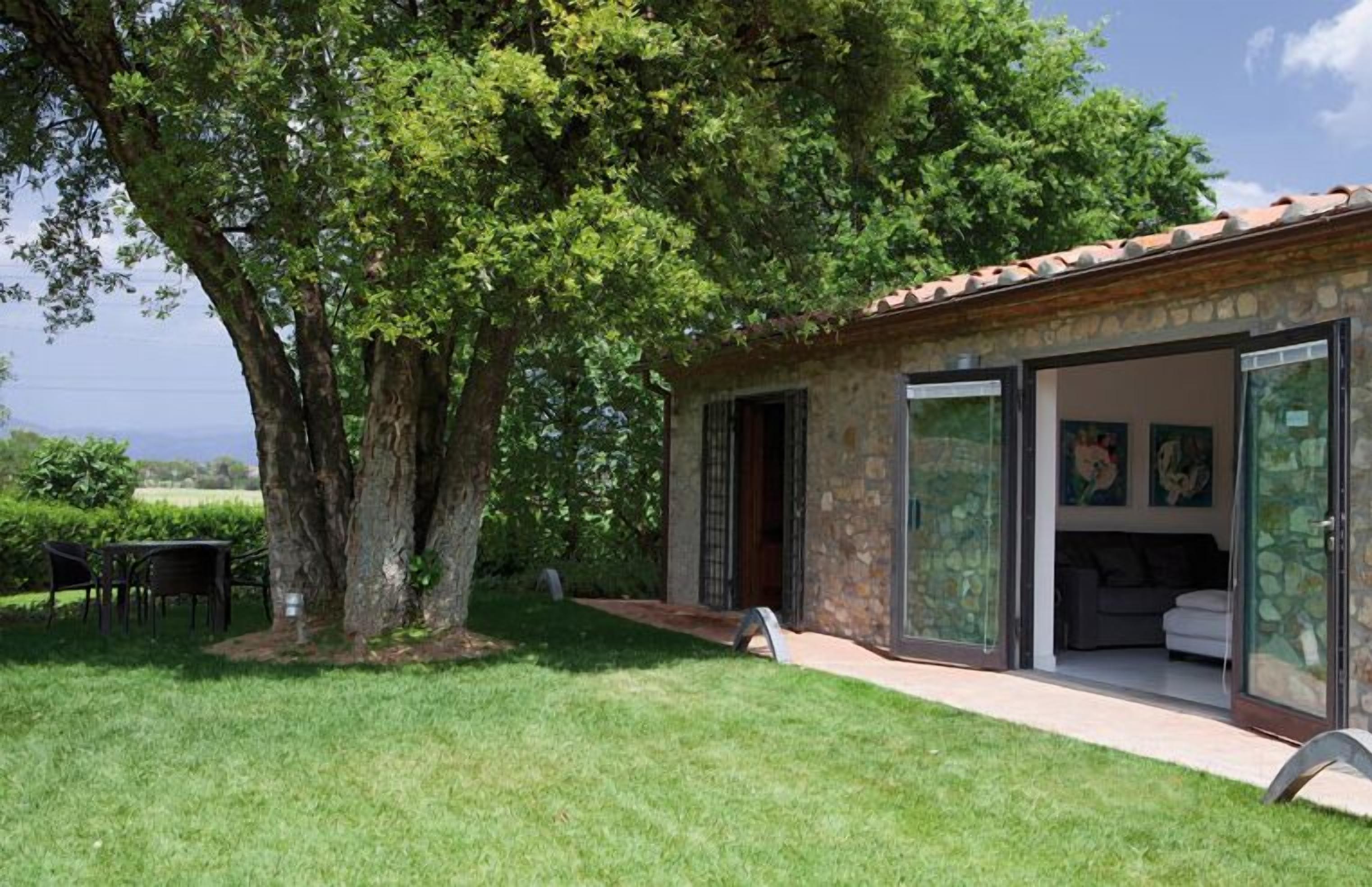 Un edificio de piedra con tejado de tejas, un cristal que da a una sala de estar, y un gran árbol en primer plano.