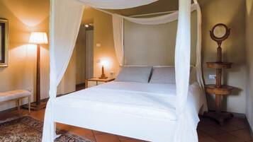 Una cama con dosel y cortinas, una mesita de noche con lámpara, y un espejo sobre un atril de madera.