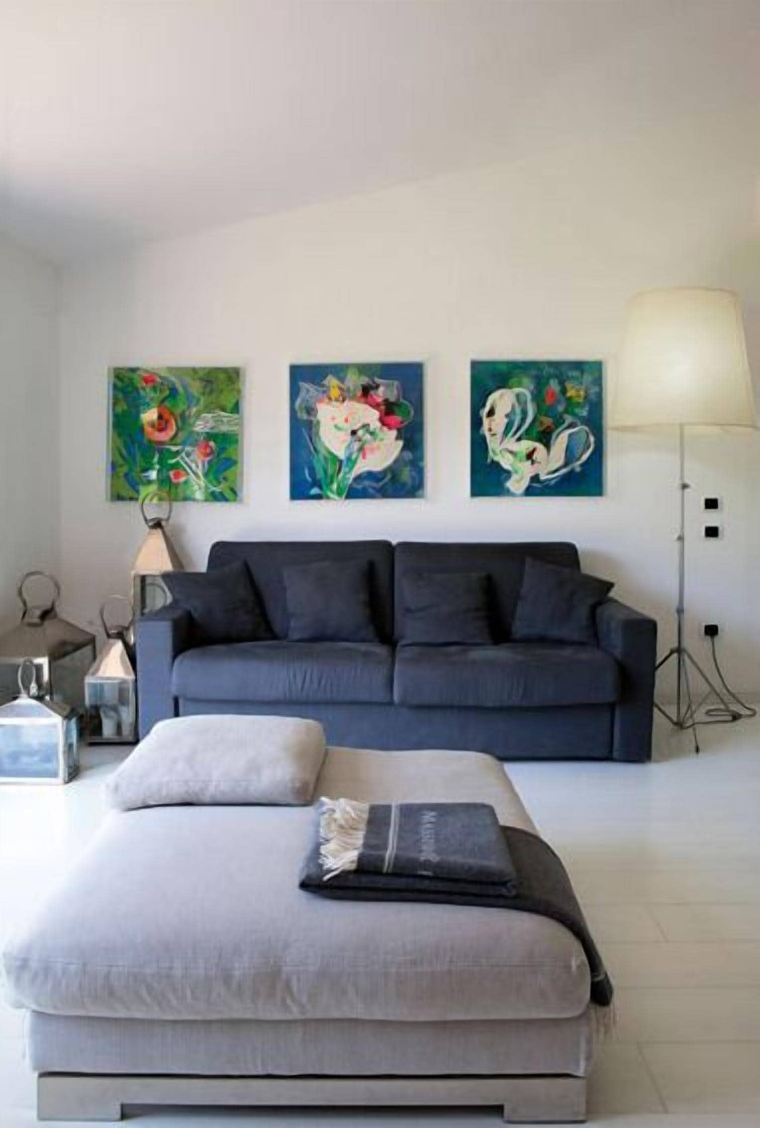 A modern living room with a grey sofa, a grey ottoman, and three colorful paintings on the wall.